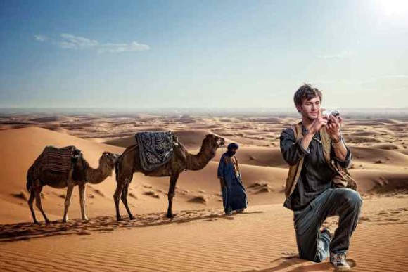 Mann kniet in der Wüste vor Kamelen mit einer Kamera in der Hand 