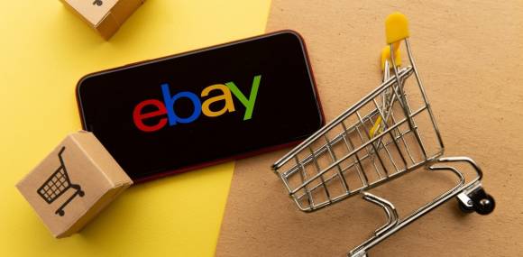 Ebay Logo auf Smartphone Screen, daneben Pakete und Einkaufswagen 