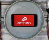 Delivery Hero App aus Smartphone, im Hintergrund Teller und Besteck