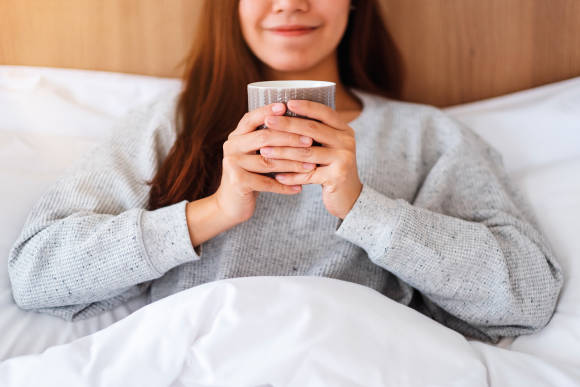 Eine Frau im grauen Pullover sitzt im Bett und trinkt aus einer Tasse 